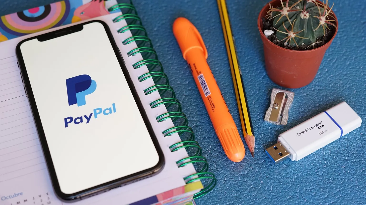 Paypal App on iPhone Screen on Opened Agenda Book with Stationery on a Blue Table