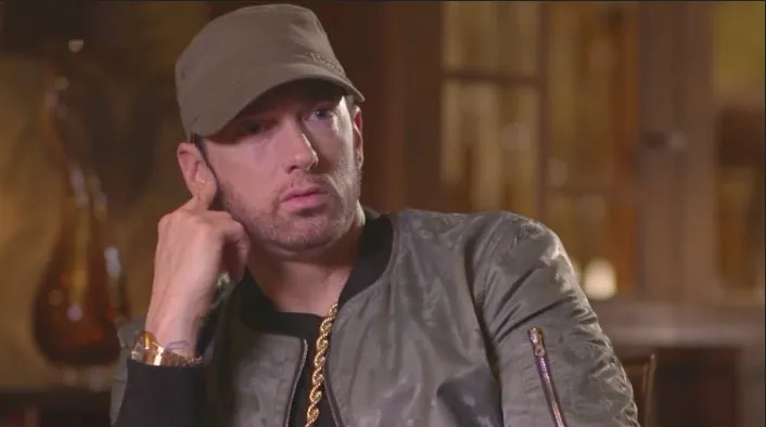 Eminem lleva un sombrero de color verde militar, una chaqueta y una cadena de oro, mientras muestra una expresión seria.