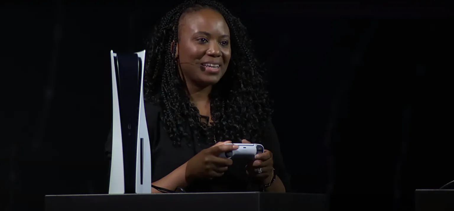 Captura de pantalla que muestra a una mujer sosteniendo un controlador de PS5 con la consola PS5 junto a ella. Fondo negro.