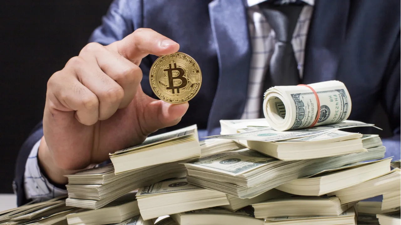 man holding a bitcoin atop a pile of US dollars