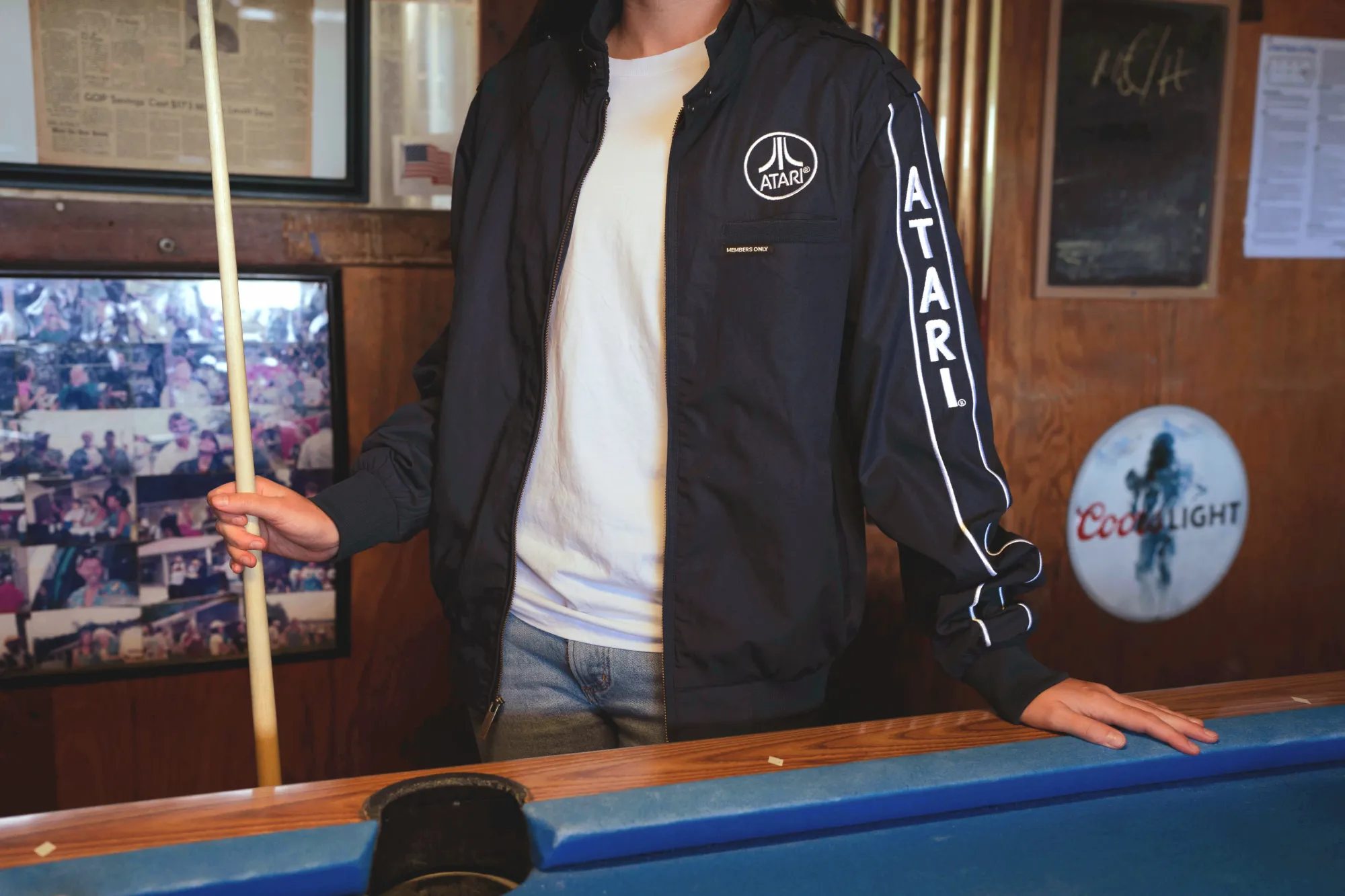 Photograph of man from neck down wearing navy and silver Atari jacket unzipped with white tshirt underneath. He holds a pool cue and has one hand resting on a blue pool table.